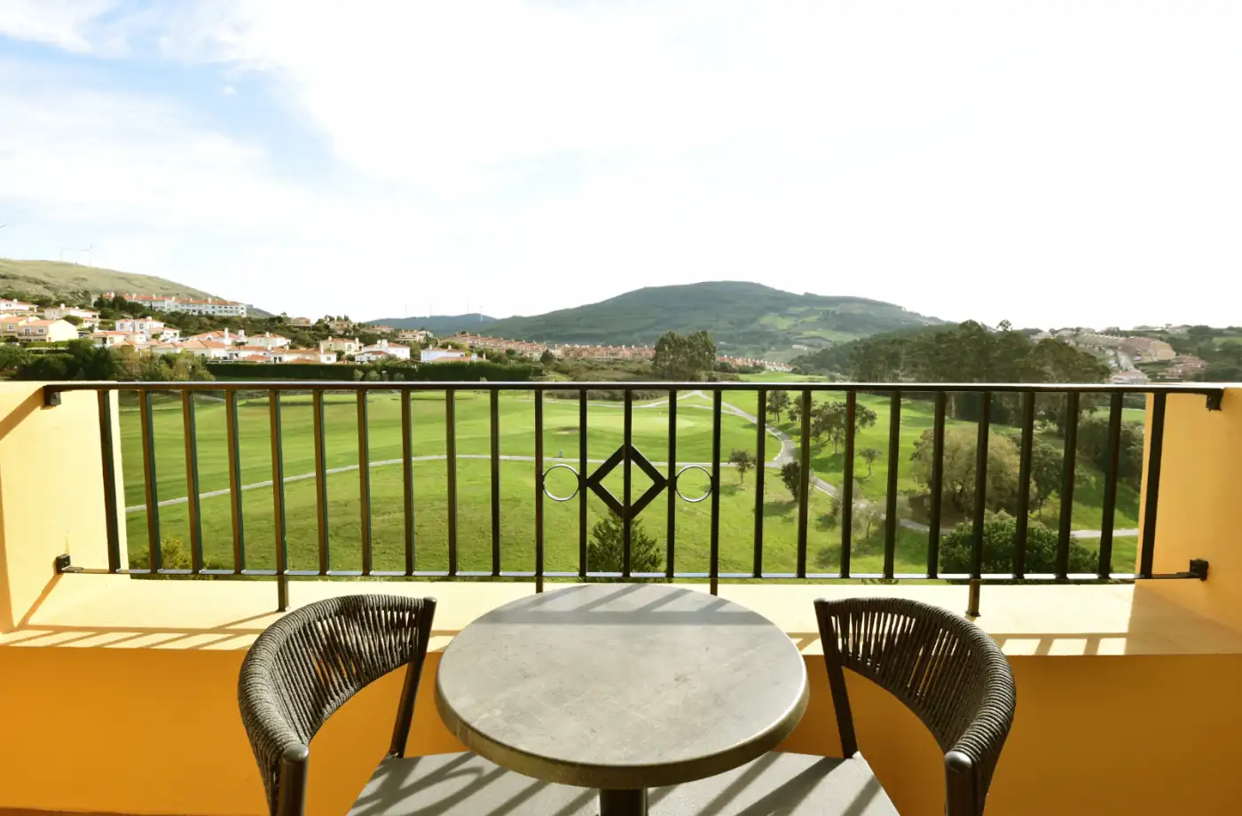 Balkon im Hotel mit runden Tisch und Stühlen, Blick auf die grüne Landschaft und Hügel im Hintergrund.