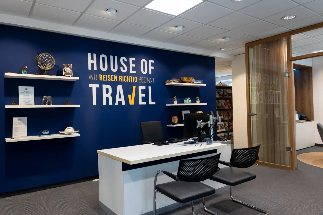 Das moderne Büro des House of Travel mit einem Empfangstisch, weiteren Sitzgelegenheiten und einer blauen Wand lädt zur Reiseberatung ein.