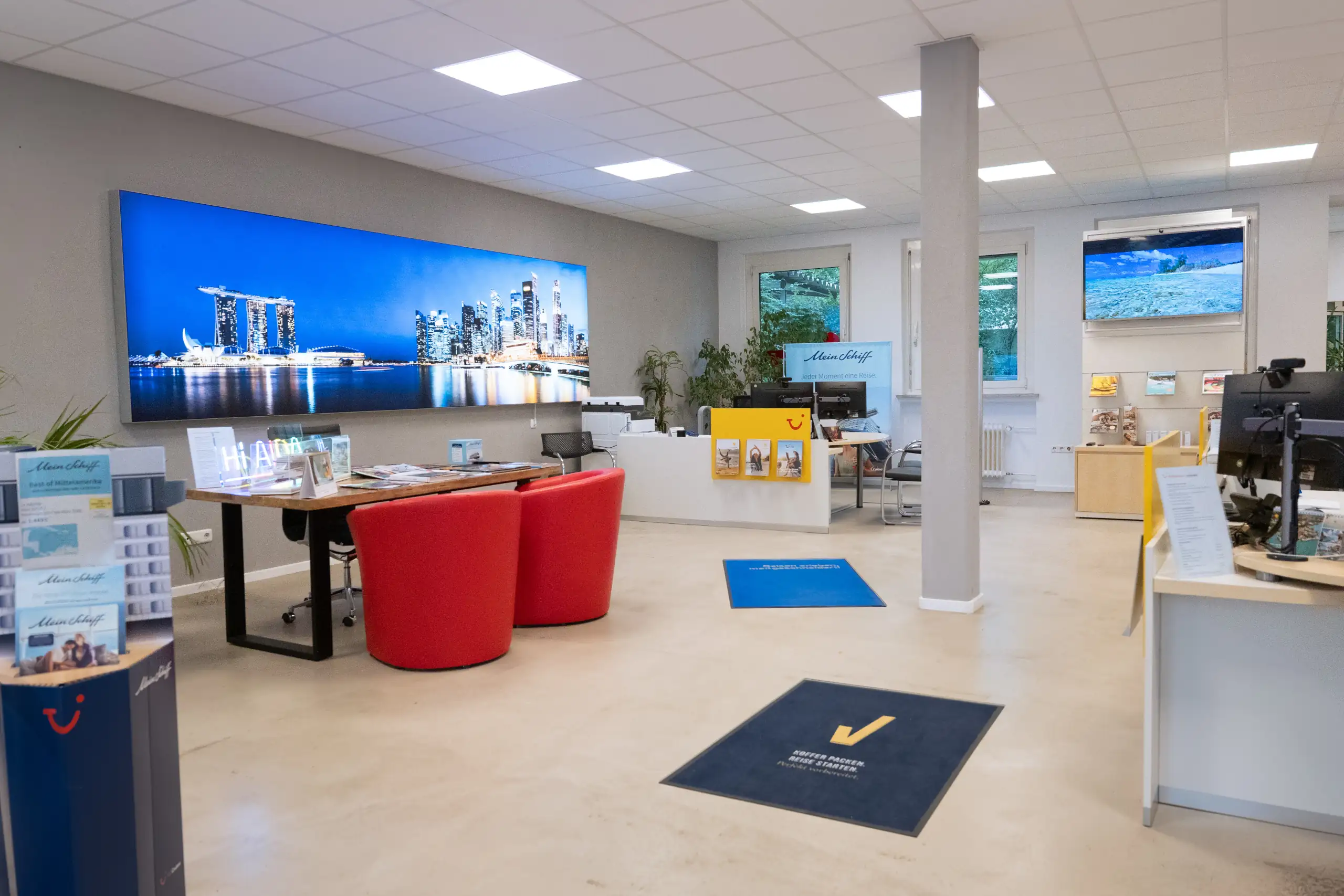 Moderne Reisewelt bei Mein Schiff – Ihr Traumurlaub beginnt hier. Blick in das moderne Reisebüro von Mein Schiff mit einem großen Stadtbild, freundlichen Arbeitsbereichen und Informationsmaterialien für die Kunden.